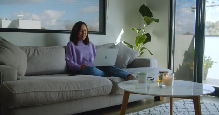 A woman sits at home and waves at her laptop as she video chats