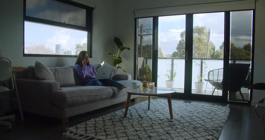A woman on a smart phone sitting on a couch next to a table with a window behind her