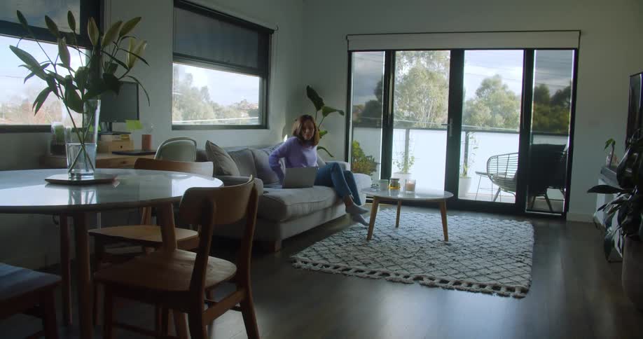 Woman walks into living room setting down drinks on coffee table before sitting on couch with laptop