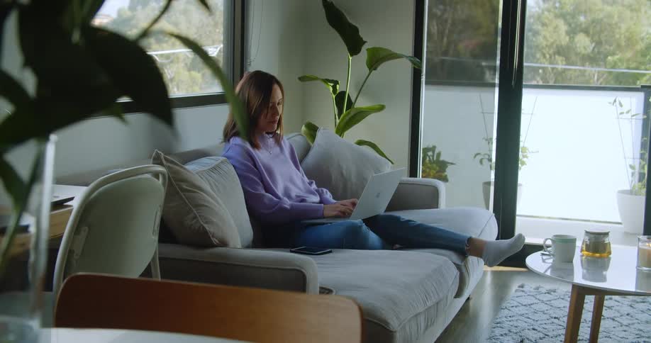 Woman sitting on couch in living room typing on laptop