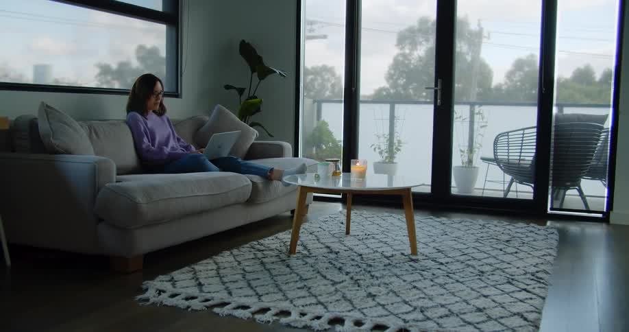 A woman sits on her couch at home and types on her laptop