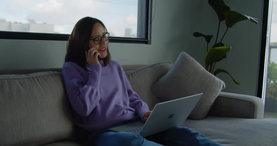 A woman sitting on a large soft couch pauses from typing on her laptop to answer a call on her smartphone