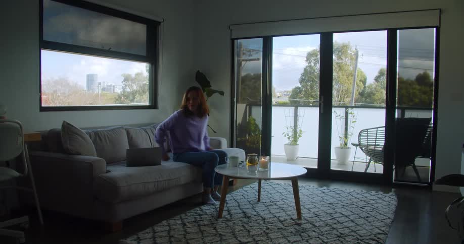 A woman carrying a coffee cup and a pot sits them on a table before sitting on a sofa and lifting a laptop