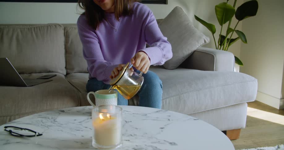 A woman sitting at home on her couch with her laptop beside her makes herself a cup of tea