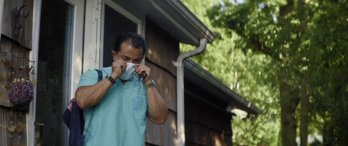 A man in medical scrubs stands on his house porch and puts a mask on and then locks the door