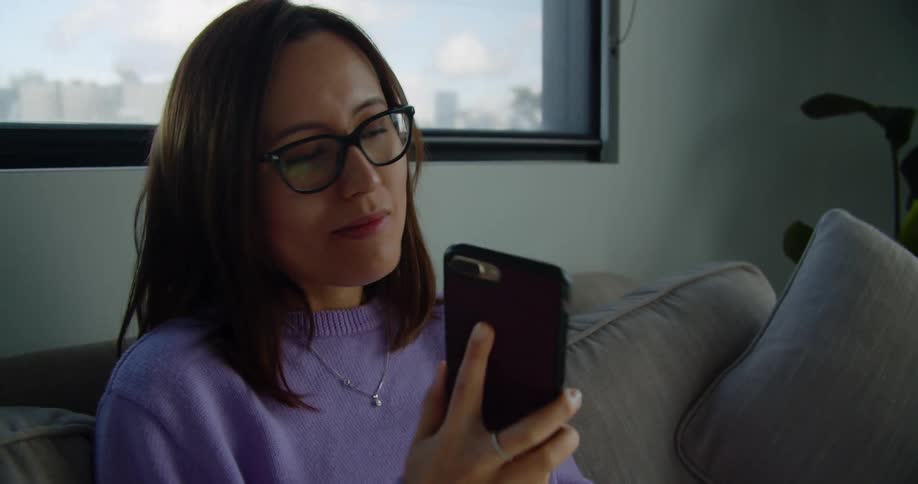 A woman answers her smartphone while sitting on a gray sofa in front of a window