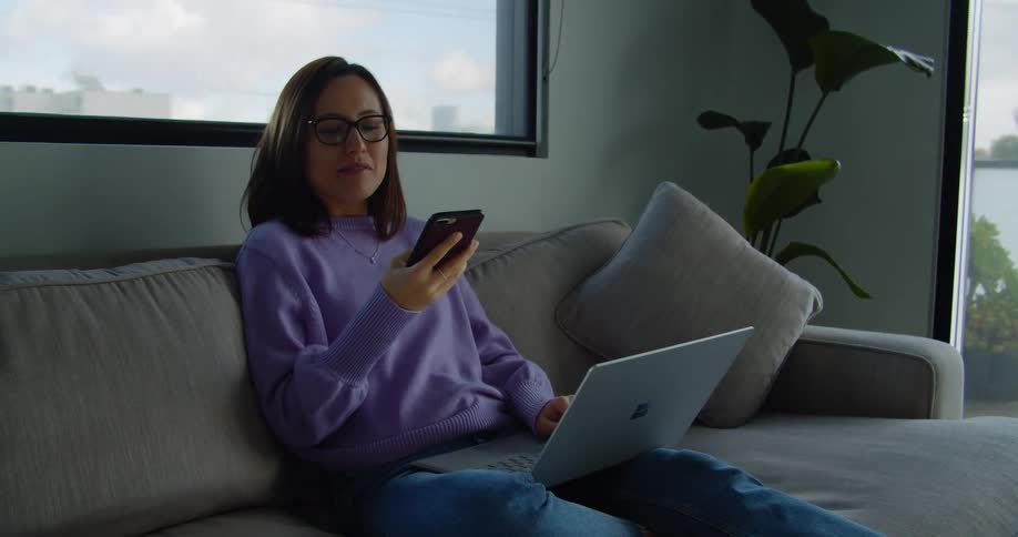 A woman talking on a smart phone with a laptop in front of her sitting on a couch