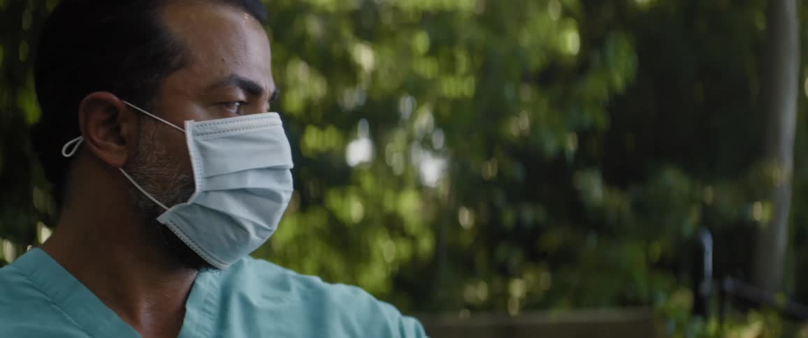 Man wearing scrubs looking toward vegetation takes off medical mask