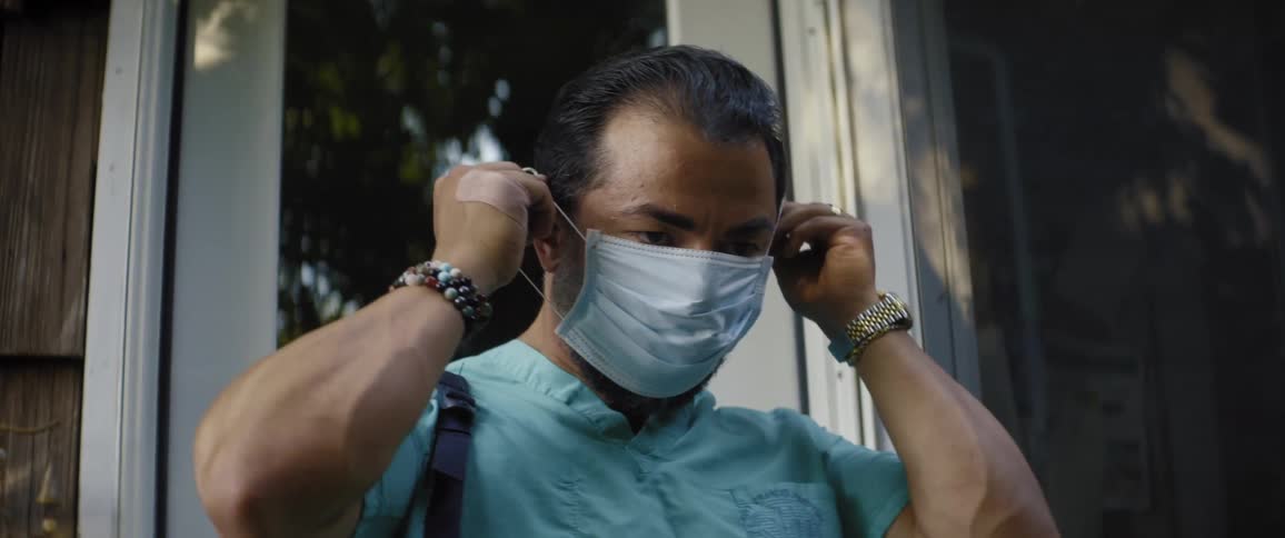 A man wearing surgical scrubs and carrying a satchel exits as house and immediately dons a disposable face mask
