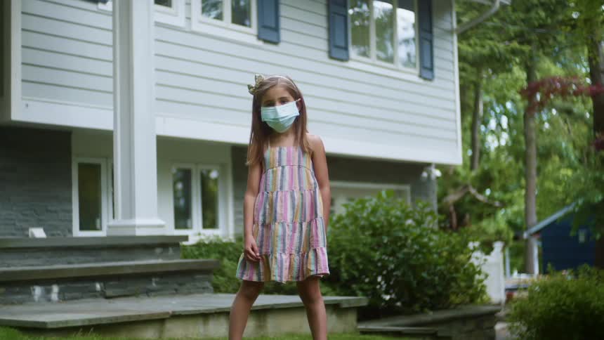 A young girl stands outside her home wearing a medical face mask