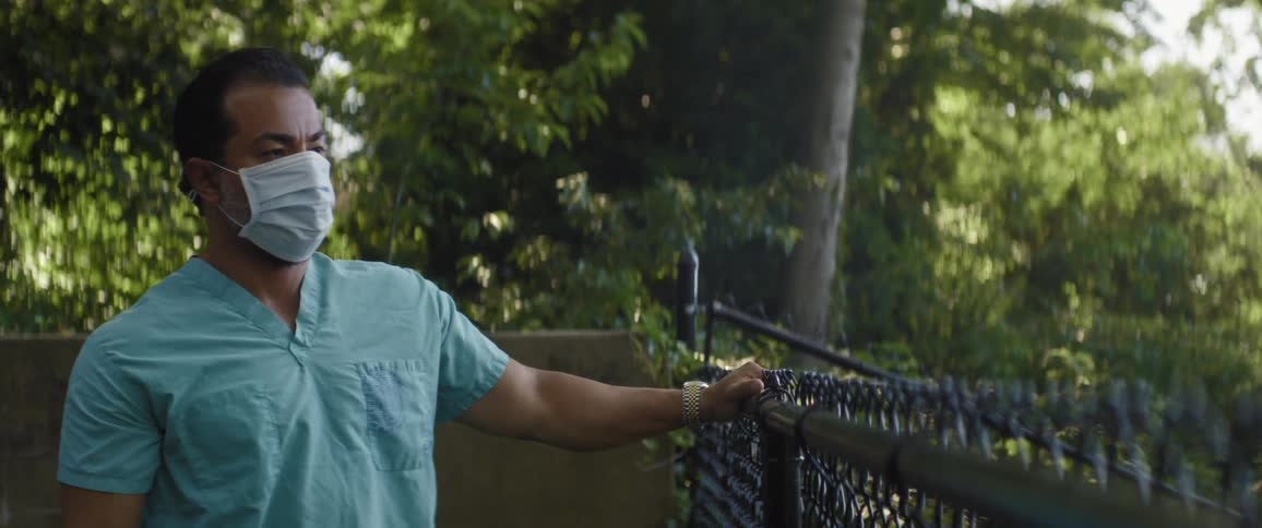 A doctor in scrubs leans against a fence outside