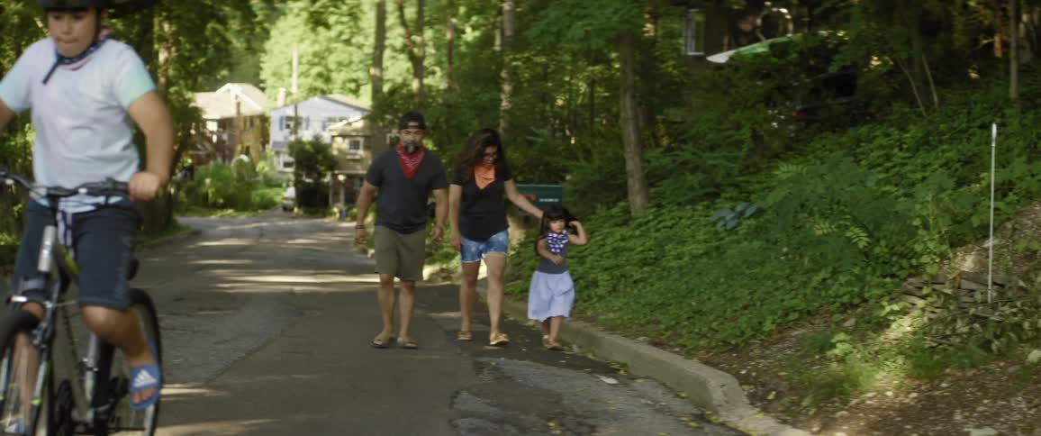 Boy rides ahead on bicycle as rest of family continues walking along road near curb