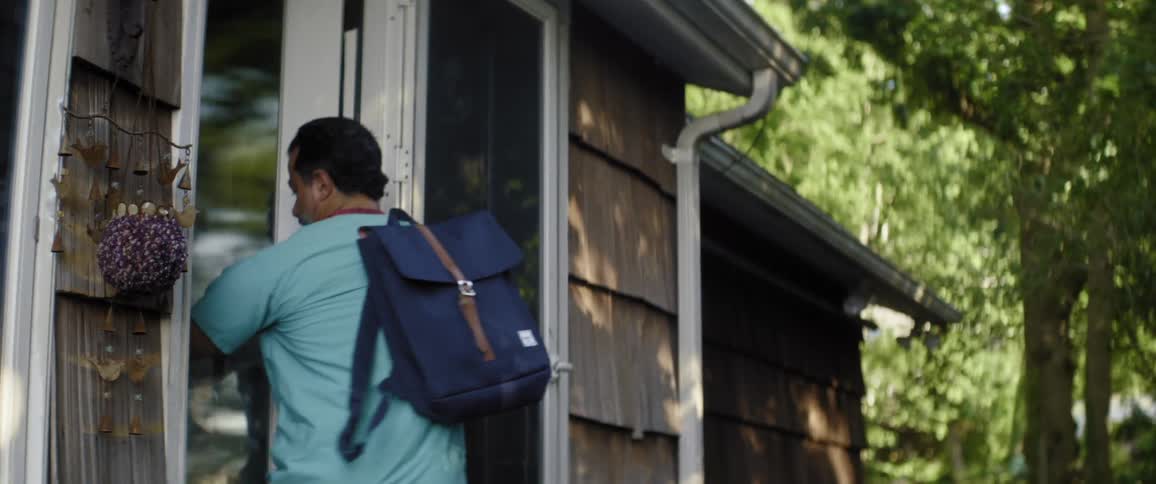 A man wearing scrubs steps out his front door and pauses to put his shoes on