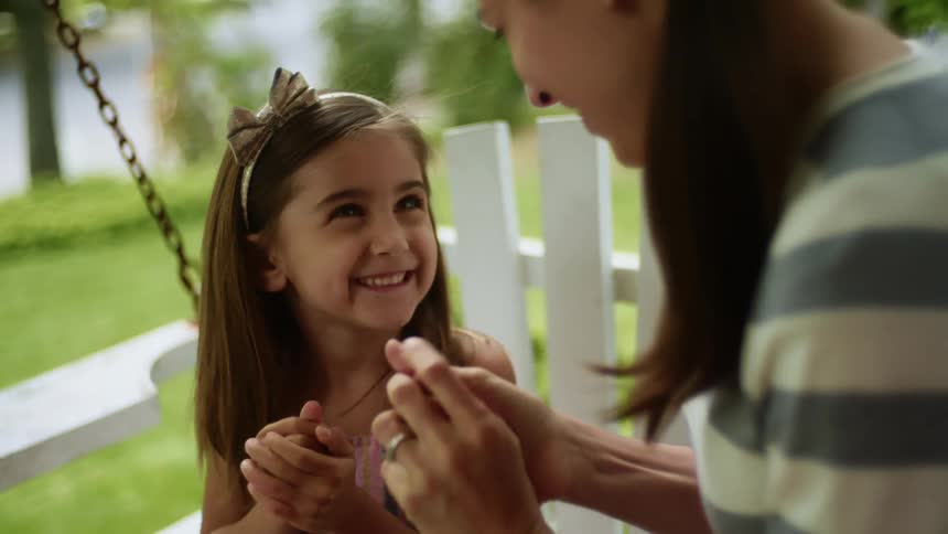 A little girl and her mom sit outside on a swing and rub their hands together and laugh