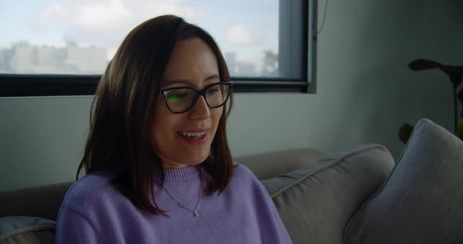 A smiling woman sitting on a couch waves as she talks to someone via video chat