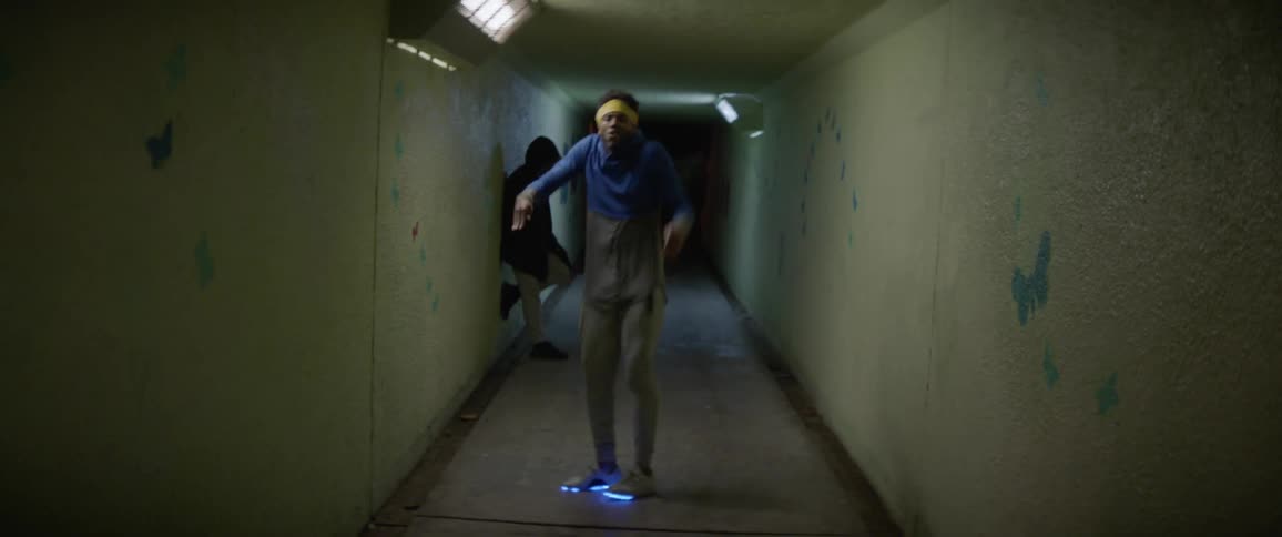 A man dances in a hallway at night while another stands leaning against the wall