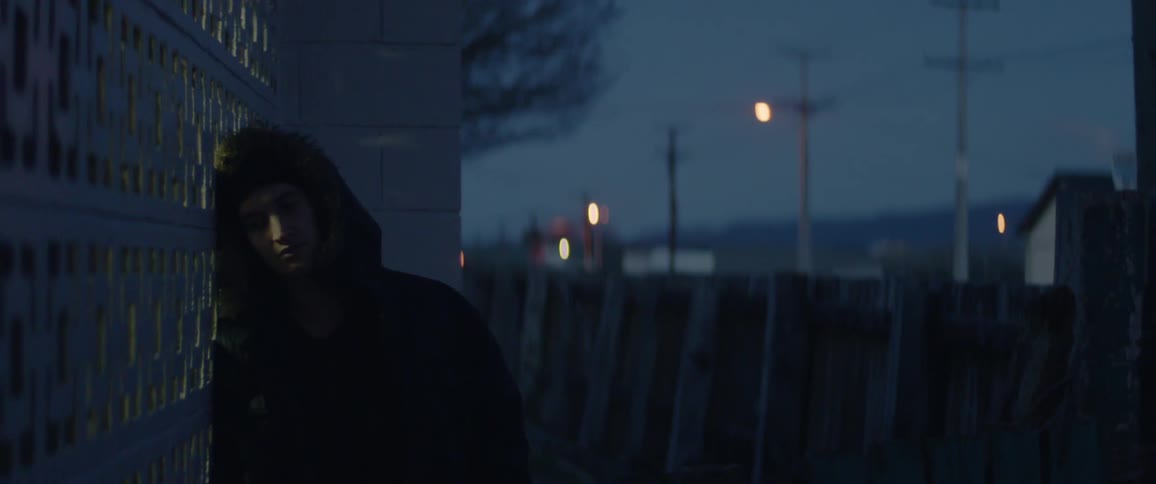 Hooded person leaning against a wall in a dark area of a city