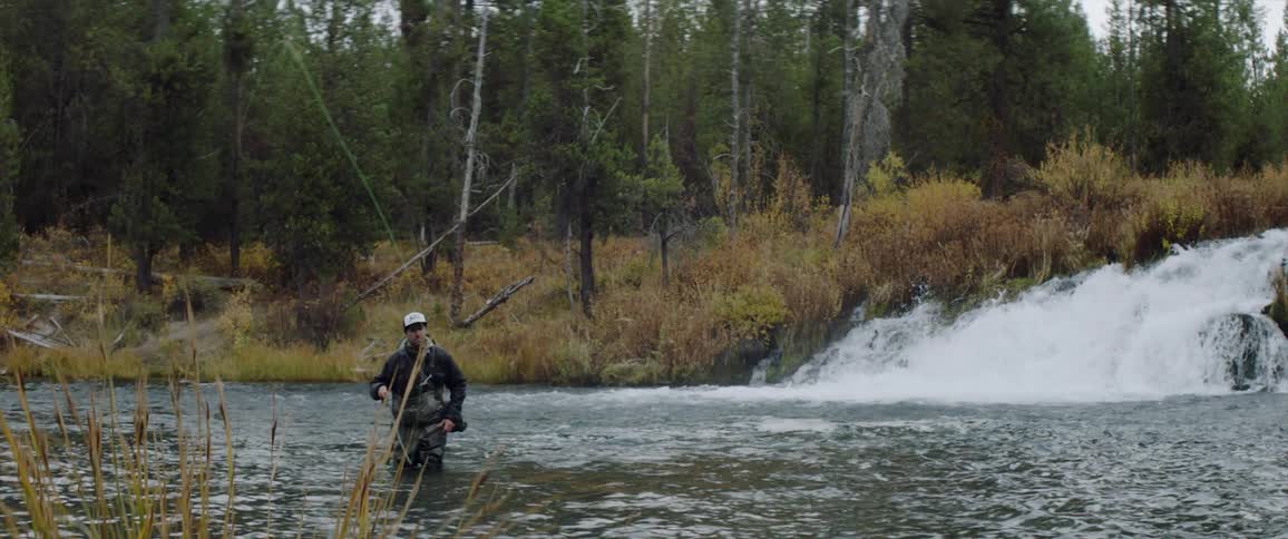 Man is fly fishing in a river near a small waterfall