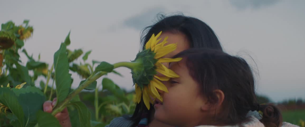 A mom smiles as she lets her daughter smell a sunflower in a field