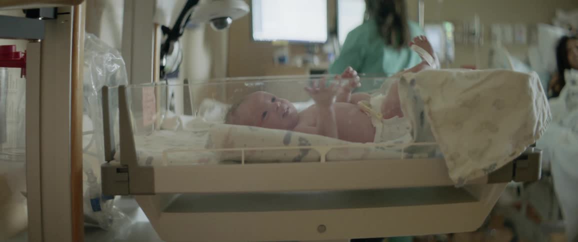 A newborn in a hospital bassinet while a nurse looks at a screen