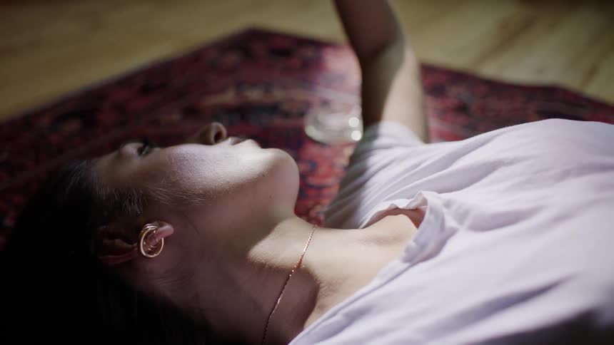 A woman lays on the floor on a persian rug, playing with the sunlight, blocking it out of her eyes with her hand.