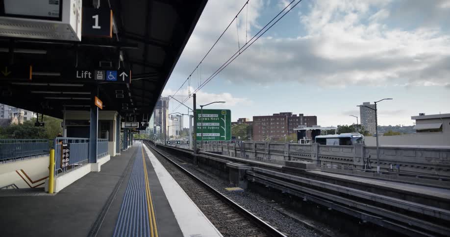 A train platform with train tracks with buildings the sky and clouds in the background