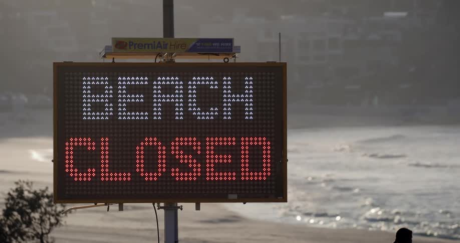 A lit sign states the beach is closed as people walk toward the waves and cars drive past