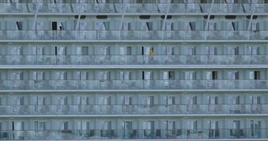 A few people stand on their balconies in a large building with several floors of identical rooms