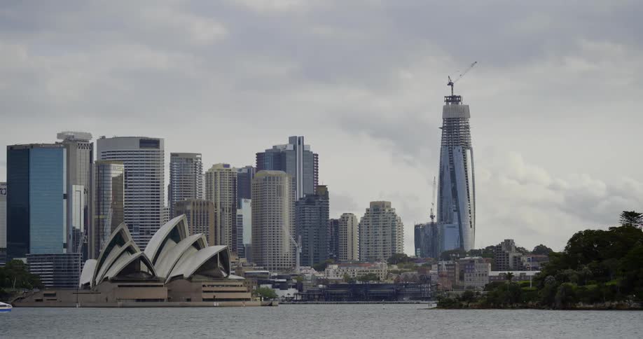 Boat cruises by sydney opera house toward tree covered shore