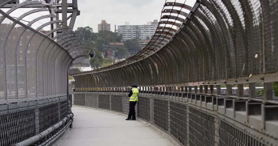 A police officer in yellow vest leans against the rail of a pedestrian bridge as cars travel on the street beside