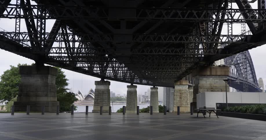 Under a bridge with a bench and pillars with an opera house and the sky in the background