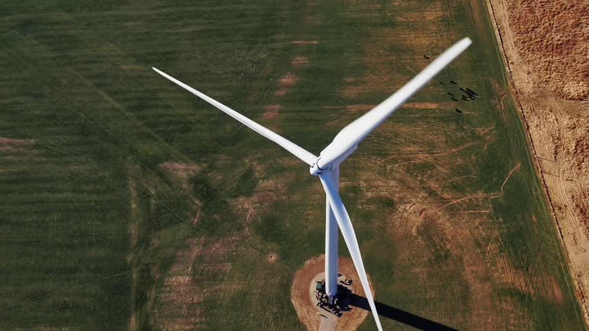 White wind turbine spins slowly casting shadow across green field and nearby bare ground