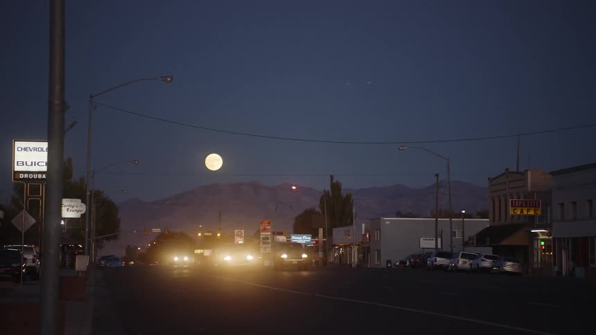 In a small town there are a handful of cars driving along a road lined with commercial buildings