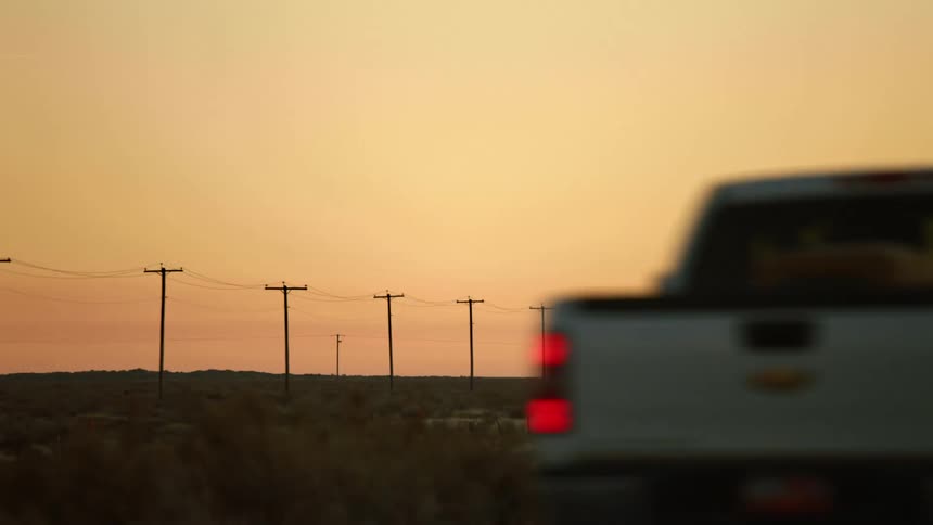 A road with a truck driving by with a grassy plain utility poles the sky and clouds in the background