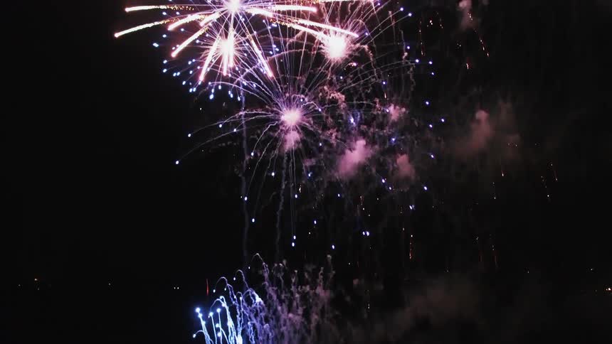 Fireworks launch from boat floating on water near city forming display in sky