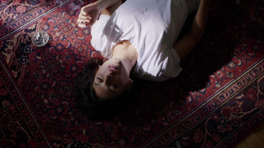 A woman relaxes on a red carpet smoking a cigarette as sunlight shines onto her from a window