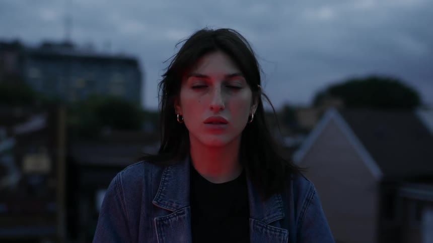 A woman stands outdoors at dusk, looking into camera with a red glow on her face.