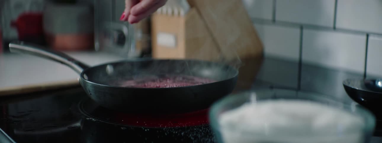 Womans hand takes some salt from a glass salt cellar and adds to the cooking food on the stove