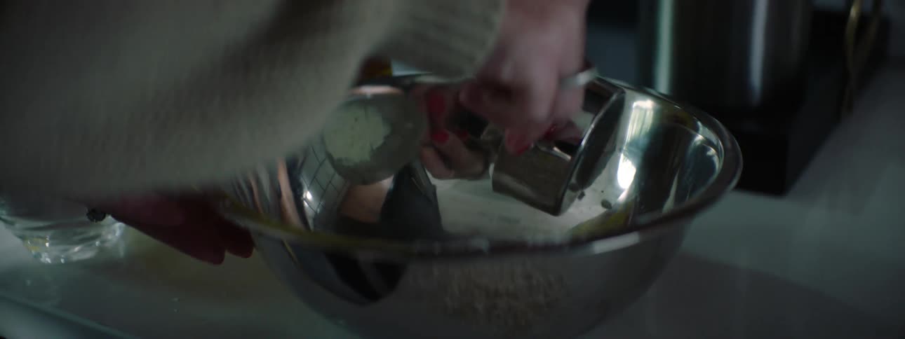 A woman measures out a powdery mixture in a metal measuring cup before pouring it into a metal mixing bowl