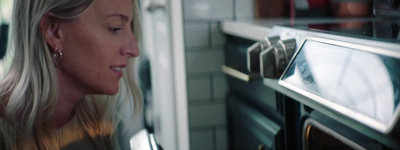 Blonde woman with earrings and a white sweater checks muffins in the electric oven in the kitchen