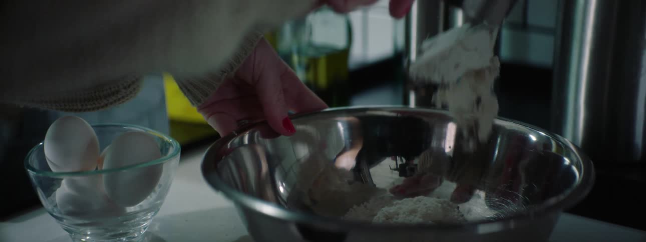 A woman scoops a cup of flour into a metal mixing bowl in a kitchen