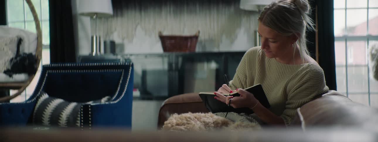 A woman sitting on a couch next to a dog writing in a book with a pen