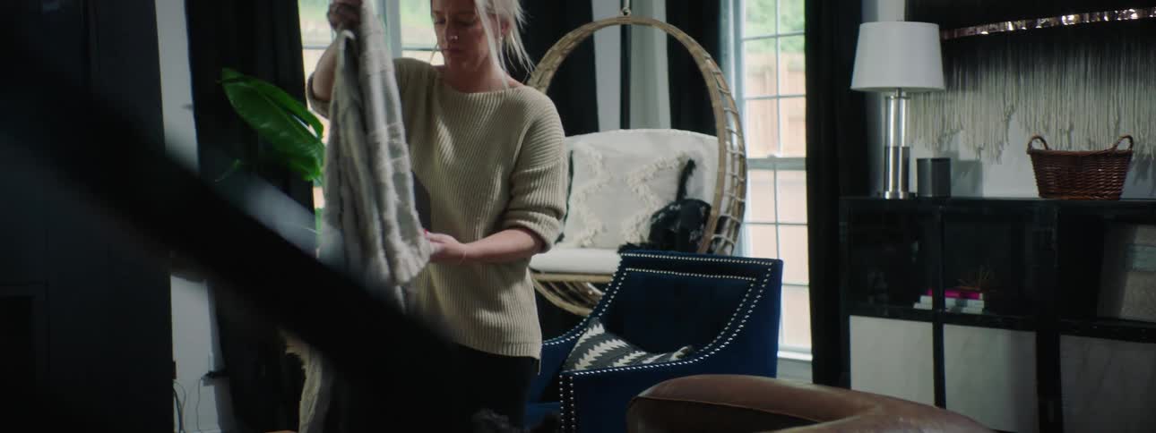 A young woman walks into her living room and sits down on the couch with a blanket and her journal