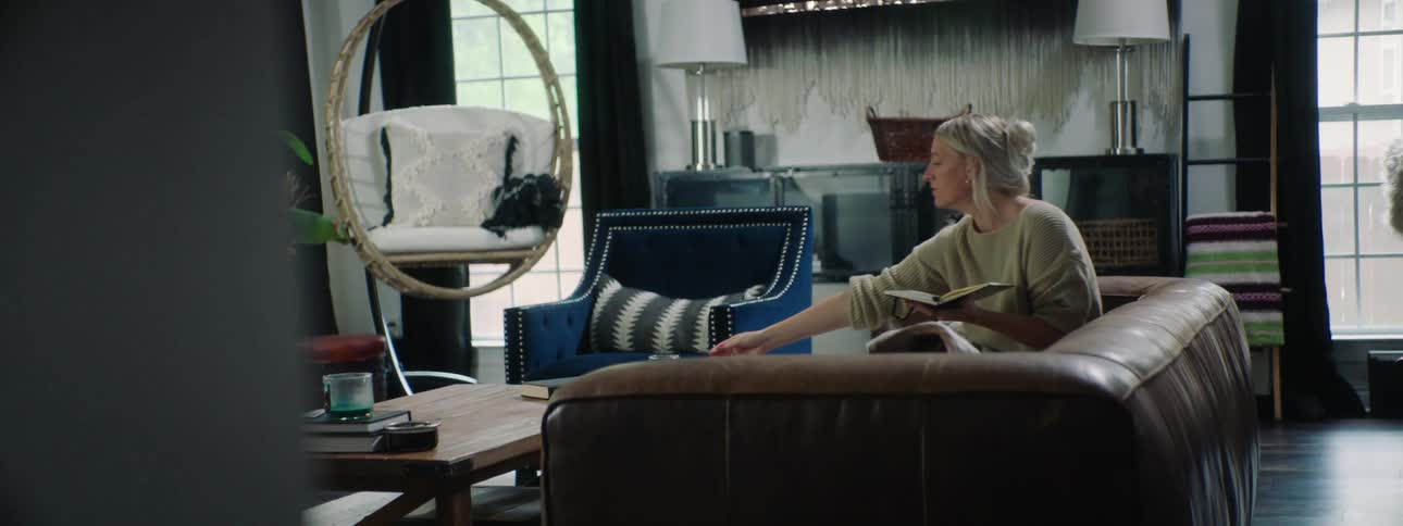 Blonde woman wearing a white sweater sitting on a brown sofa writes in a notebook and drinks a cup of tea in the living room