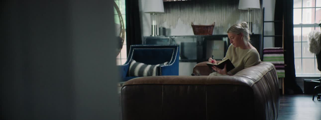 Blonde woman wearing a white sweater sitting on a brown sofa writes in a notebook and a small dog lifts its head in the living room