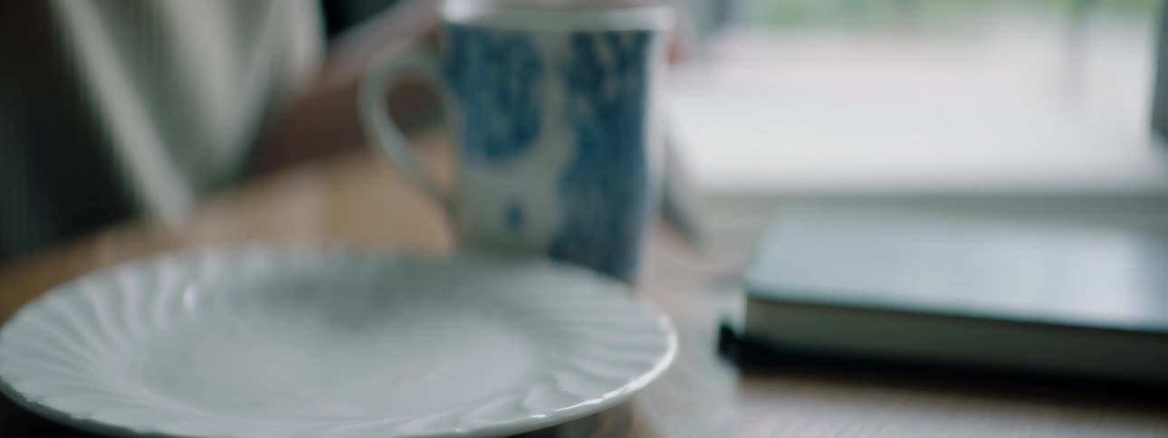 A woman sitting at a table taking a pastry off a plate next to a mug