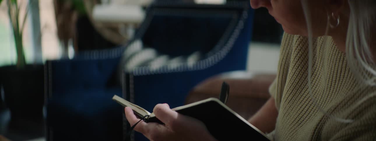 A woman writes in a notebook with a pen while sitting indoors