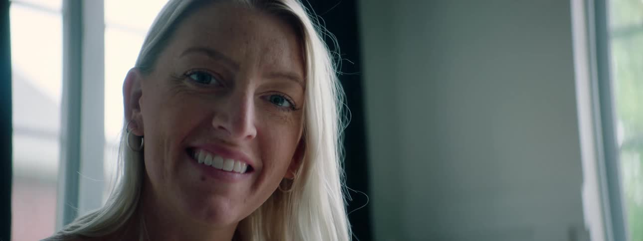 Blonde woman with earrings looks at the camera and smiles happily in a room