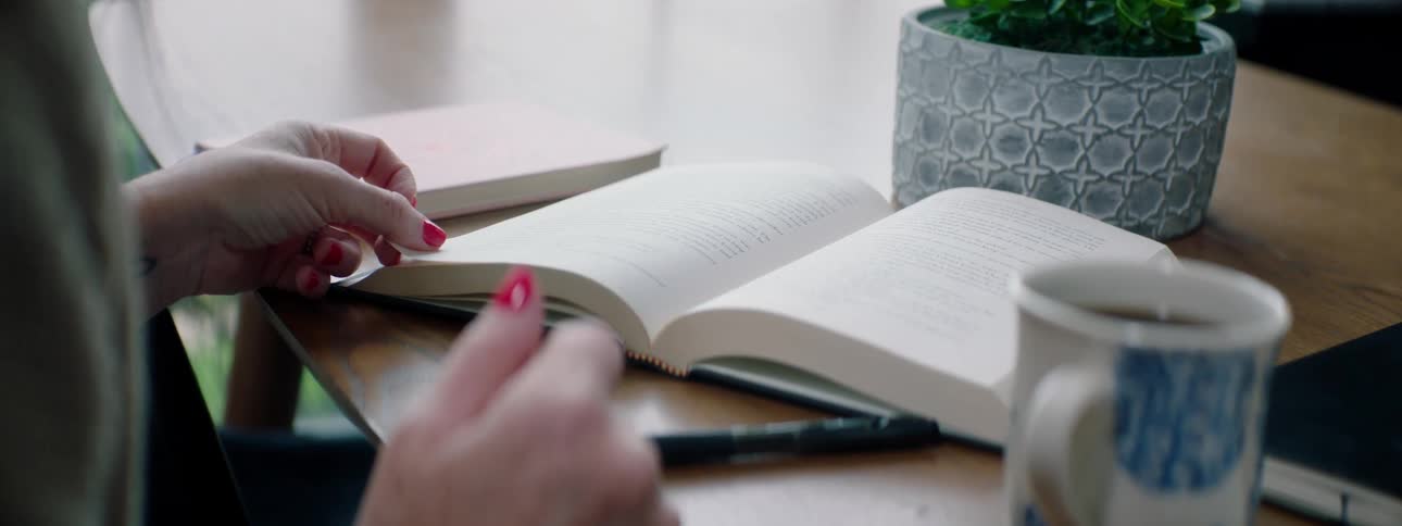 A woman sitting at a table reading a book next to a coffee cup and a plant