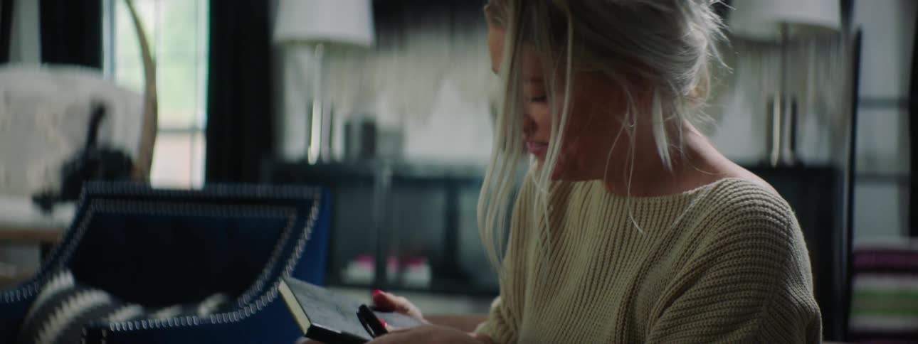 A woman sitting on a couch covering up with a blanket opening a book and petting her dogs